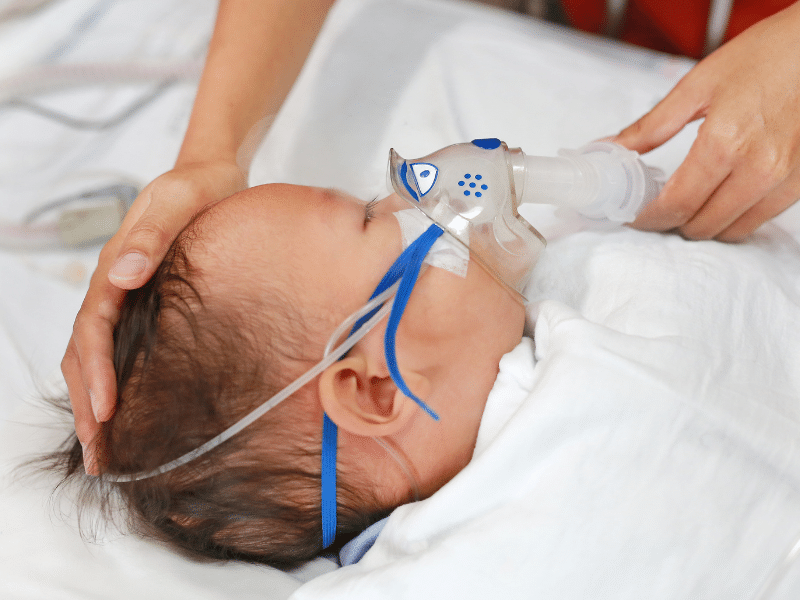 Infant wearing an oxygen mask with a caregiver’s hands gently supporting the baby during breathing treatment.